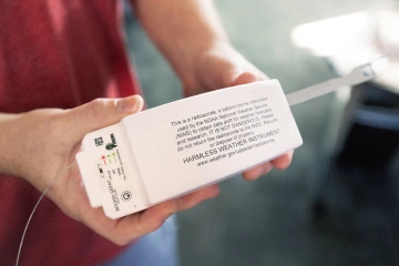 A man in a red shirt holding up the radiosonde to the camera. Writing is seen on the radiosonde. The writing explains what the radiosonde is, who is belongs to and states it is harmless.