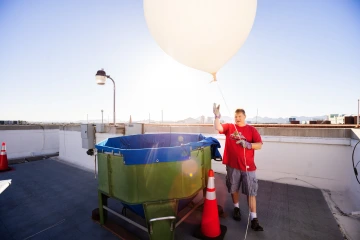 A man in a red Arizona Wildcats shirt holds the string of a large white weather balloon on a rooftop, preparing for its launch. A green tank with a blue lining and a safety cone sit nearby, with the sun shining brightly in the background.
