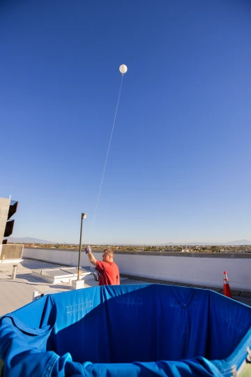 A man in a red shirt releases a weather balloon, which ascends into a clear blue sky, trailing a thin string. A large blue-lined tank is visible in the foreground, with city rooftops and mountains in the distance.