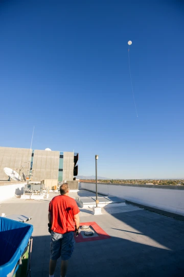A man in a red shirt watches a white weather balloon as it rises high into the bright blue sky above a rooftop filled with satellite dishes and equipment, with distant mountains in the background.