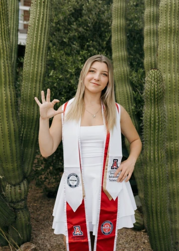 An image of Arizona Alumni Aurora Seekins at her graduation photoshoot. She is seen holding up the Wildcat hand sign. Cacti are visible in the background.