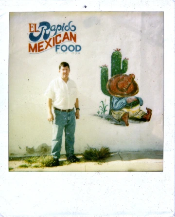 A man standing next to a mural of a man wearing a sombrero next to a cactus with the "El Rapido Mexican Food" slogan.