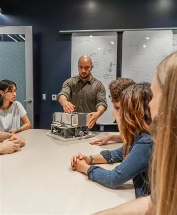 Principal Investigator Carlos Vargas standing at a table with seated students, surveying a 3D-printed  model of the Aspera telescope in the Applied Research Building. 