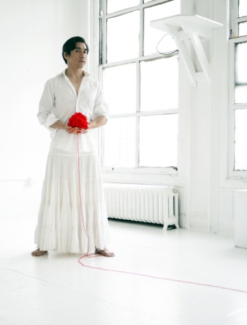 A photograph of Joseph Legaspi, dressed in white, in a white room, holding a red yarn ball, which has left a train of red yarn
