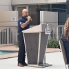A man in a navy blue shirt speaking into a microphone at a podium. The podium is positioned near a swimming pool and features the University of Arizona logo with the word "ARIZONA" on it. In the background, there is a railing and plants adjacent to a modern building with large windows.
