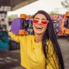 A smiling young woman with long black hair and red-tinted cat-eye sunglasses holds a colorful skateboard over her shoulders. She’s wearing a bright yellow sweatshirt and standing in an urban skate park with graffiti-covered walls. The skateboard has a vivid, artistic design with bold colors and orange wheels.
