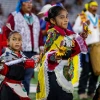 Two young children in traditional Hopi regalia perform a dance on the field, holding corn and flowers, during the Hopi Recognition Football Game at Arizona Stadium, with adult participants standing behind them.