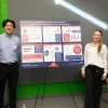 A group of four individuals stands in front of a large poster titled "What Drives Employment Success?" The poster includes charts and bullet points about education, income, and job stress. The group smiles at the camera, wearing business casual attire, with a green and gray wall in the background.