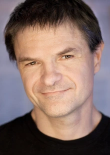 A close-up portrait of a smiling man in a black shirt against a softly lit, out-of-focus background.